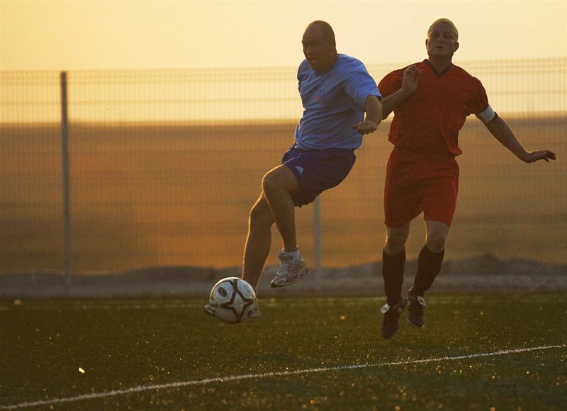 Two players chasing a soccer ball across a grass field at sunset